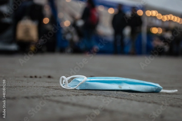 Fototapeta Close up of a blue and withe mouth mask lying dirty on the street in corona covid time. People and lights in the background