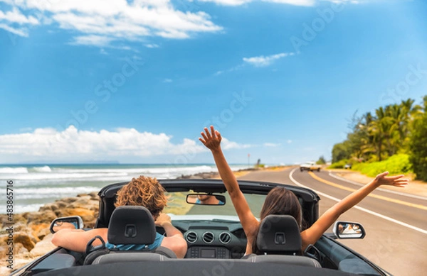 Fototapeta Road trip car holiday happy couple driving convertible car on summer travel Hawaii vacation. Woman with arms up having fun, young man driver