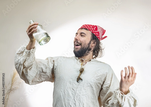 Fototapeta Portrait of a young dark-haired man with a beard with a red cloth bandana on his head and an old shirt with a bottle of rum on a light background. Pirate Costume