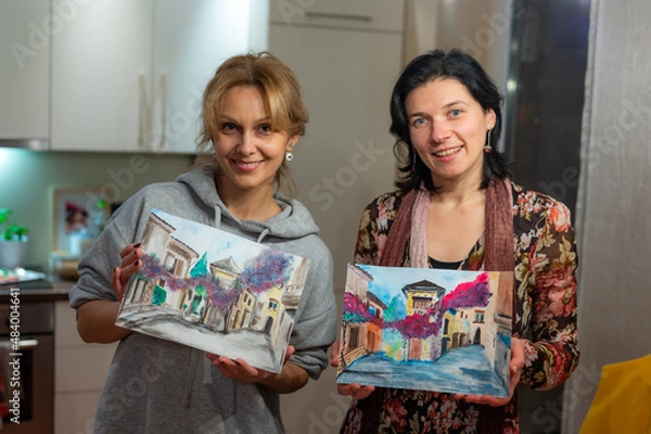 Fototapeta Two happy young women holding finished watercolor paintings of vibrant medieval european cityscape in their hands.