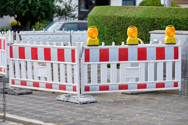 Obraz Street barriers with yellow warning lights standing in front of a construction site on the pavement in front of a green hedge. Seen in Germany in October.