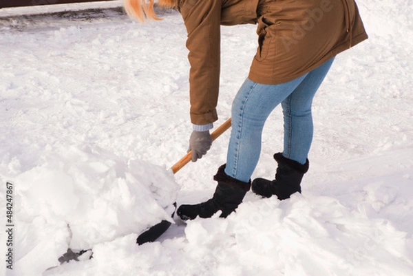 Fototapeta Snow removal in winter. A woman cleans the snow with a shovel in the local area. Woman clearing snow from her front yard.