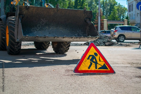 Fototapeta close up, a sign, a symbol of road works is installed on the asphalt against the background of a city street with a blocked passage and a tractor, a bulldozer with a raised bucket