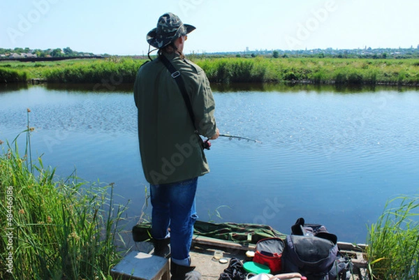 Fototapeta A fisherman with a fishing rod stands on a pier on the banks of the river, rear view on a sunny summer day among the reeds