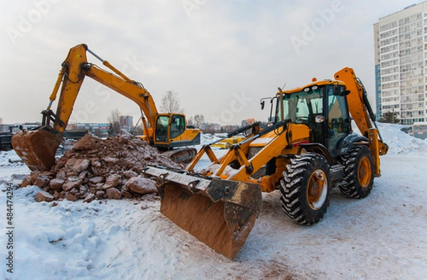 Fototapeta an excavator digs the ground at a construction site in winter against the background of a new house
