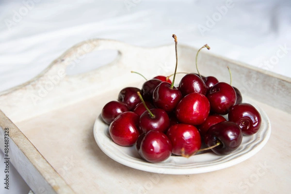Fototapeta A ripe cherry in a saucer is on a wooden tray on a light background. Fresh and beautiful cherry berries are on a plate.