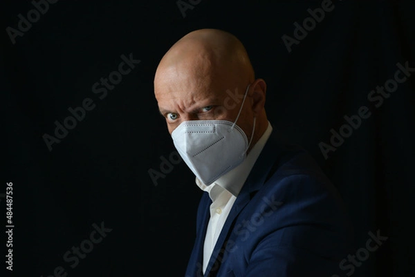 Obraz A man in a business suit with a medical mask on his face on a black background. Means of protection of respiratory organs. Precautions against infection with Covid-19 (SARS-CoV-2).