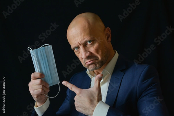Obraz A man in a business suit holds a medical mask in his hand, points to it with his index finger., black background. The man recommends the use of medical masks.