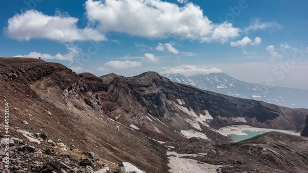 Fototapeta Acid lake on top of a volcano. Snow melting on steep slopes. The layered structure of the soil. On the edge of the crater, against the sky and clouds, tiny silhouettes of people are visible. Kamchatka