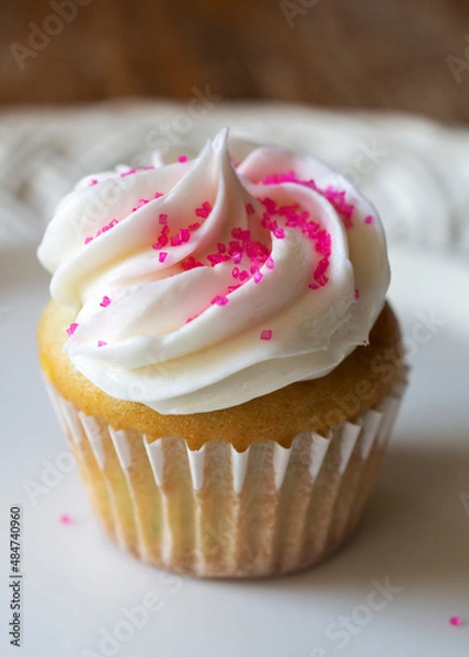 Fototapeta A close up view of a mini cupcake decorated with pink sugar sprinkles.  Confetti cupcake batter with white frosting on a white plate.  Wooden table blurred in background.