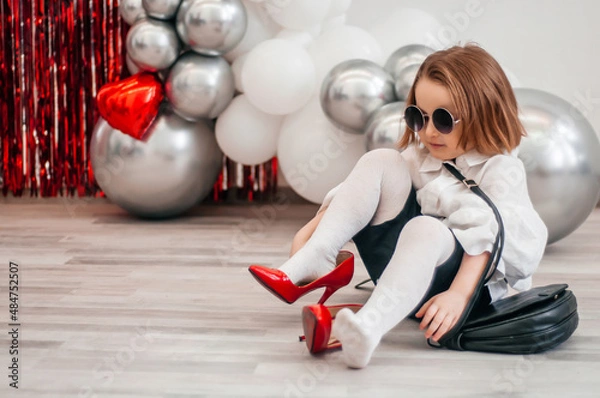 Obraz A fashionable baby in sunglasses is trying on her mom's red shoes. Fashion and modern clothes. Glitter bright metallic mirror background, new trends valentine's day
