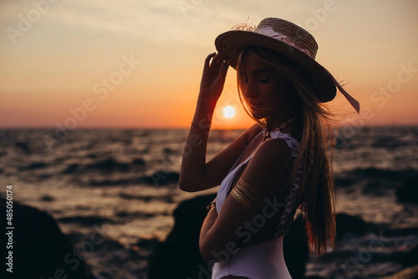 Obraz Silhouette of a girl in a hat on the background of the dawn sun close-up. Vacation, beach holiday concept.