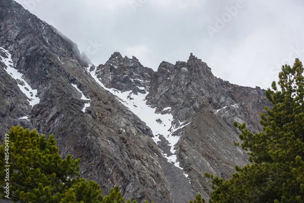 Obraz Gloomy mountain landscape with high sharp rockies with snow in gray low clouds. Closeup of sharp rocks and peaked tops in gray cloudy sky. Bleak overcast scenery with mountain range with pointy rocks.