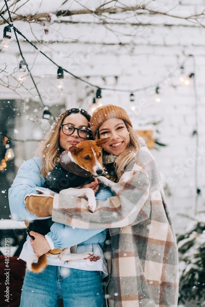 Fototapeta Portrait of two cheerful women friends standing outdoor in the backyard with dog in snowfall, enjoying winter time.