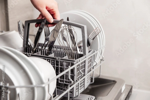 Fototapeta Close-up of female hand holding full cutlery basket with clean knife, fork, whisk. Loading to, empty out or unloading from open automatic dishwasher machine with clean utensils in home kitchen