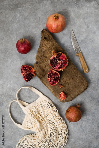 Fototapeta Beautiful red pomegranate fruit composition on a concrete background. Half pomegranate and ripe pomegranate fruit with rustic wooden board, knife and string bag. Top view.