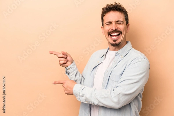Fototapeta Young caucasian man isolated on beige background pointing with forefingers to a copy space, expressing excitement and desire.