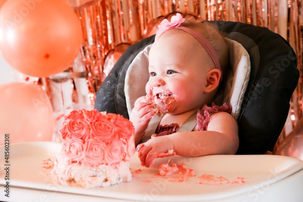 Obraz Cute birthday girl celebrating her one year birthday by messily eating a cake surrounded by pink background streamers and balloons