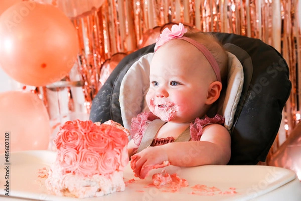Obraz Cute birthday girl celebrating her one year birthday by messily eating a cake surrounded by pink background streamers and balloons
