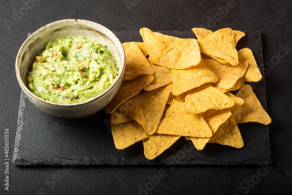 Fototapeta Mexican nachos chips and bowl with homemade fresh guacomole sauce made from avocado and lime  over black background.
