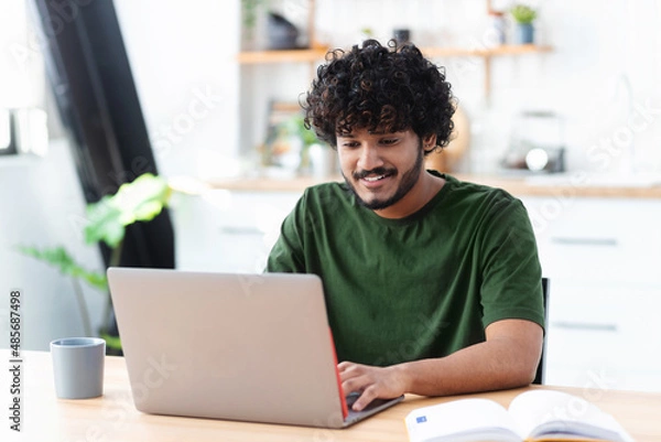 Fototapeta Smiling young indian man using laptop to work remotely from home, Portrait of successful freelancer or student working on new project
