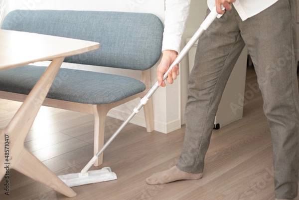 Obraz A Japenese young Asian man hoovers the floor using a vacuum cleaner in a house with a desk and two chairs
