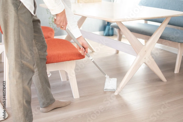 Obraz A Japenese young Asian man hoovers the floor using a vacuum cleaner in a house with a desk and two chairs