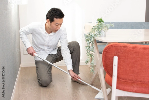 Obraz A Japenese young Asian man hoovers the floor using a vacuum cleaner in a house with a desk and two chairs