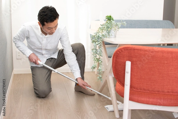 Obraz A Japenese young Asian man hoovers the floor using a vacuum cleaner in a house with a desk and two chairs