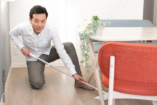 Obraz A Japenese young Asian man hoovers the floor using a vacuum cleaner in a house with a desk and two chairs