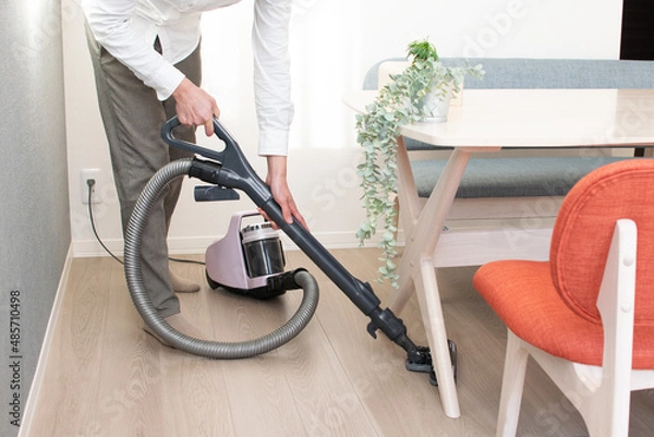 Obraz A Japenese young Asian man hoovers the floor using a vacuum cleaner in a house with a desk and two chairs