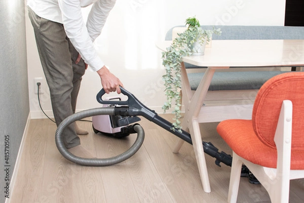Obraz A Japenese young Asian man hoovers the floor using a vacuum cleaner in a house with a desk and two chairs