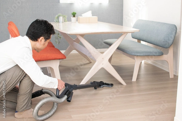 Obraz A Japenese young Asian man hoovers the floor using a vacuum cleaner in a house with a desk and two chairs