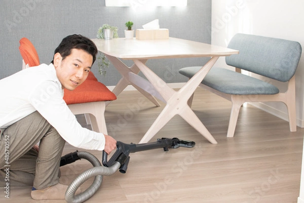 Obraz A Japenese young Asian man hoovers the floor using a vacuum cleaner in a house with a desk and two chairs