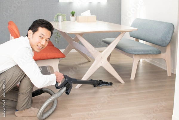 Obraz A Japenese young Asian man hoovers the floor using a vacuum cleaner in a house with a desk and two chairs