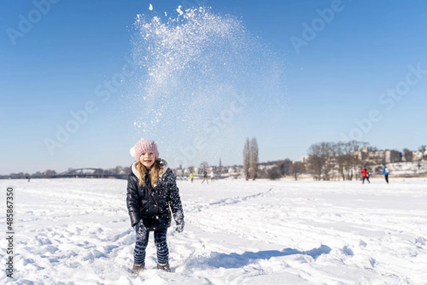 Fototapeta Young caucasian smiling preschool girl in warm clothes throws snow up playing and having fun on sunny winter weather outside on snow covered field in city park. Christmas holidays, childhood, carefree