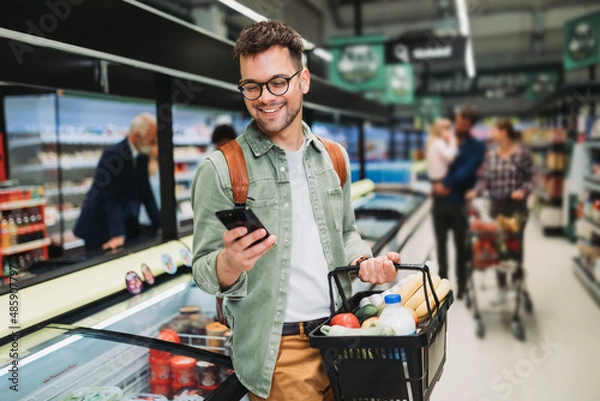Obraz Young man buying groceries at the supermarket. Other customers in background. Consumerism concept.