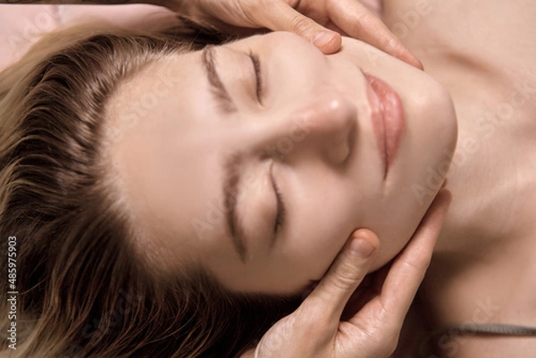 Fototapeta Young beautiful woman is getting a relaxing head and face massage in wellness center. Masseur doing massage the head of an young beautiful girl woman in the spa salon close up macro