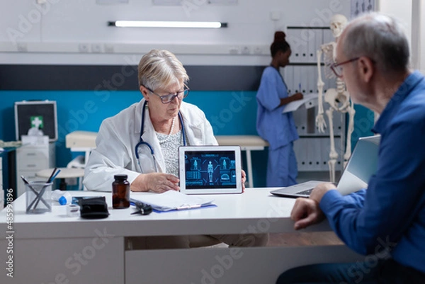 Fototapeta Medic showing tablet with illustration of human body analysis to senior man. Healthcare specialist explaining diagnosis to ill patient while holding device with virtual anatomy and expertise
