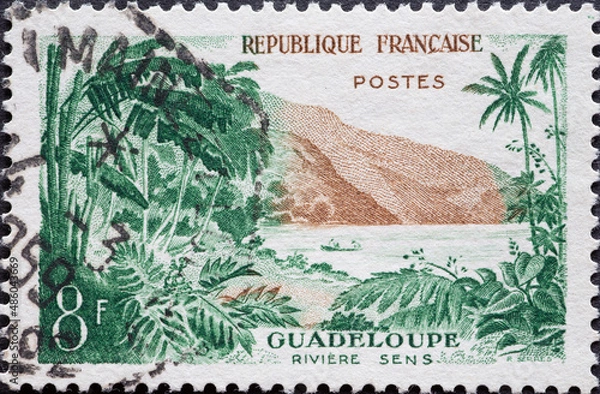 Fototapeta France - circa 1957: a postage stamp from France , showing a tropical landscape with palm trees and mountains at the Sens River, Guadeloupe
