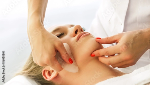 Obraz Beautician cosmetologist makes lifting modeling face massage with Gua Sha quartz scraper, professional cosmetic procedure in a beauty clinic salon for client. Skin care and cosmetology spa concept.