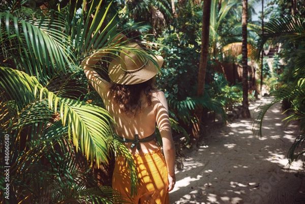 Fototapeta Young beautiful woman stands in the shade of palm trees, view from the back. A girl walks among tropical greenery on a bright sunny day in Mexico, Tulum