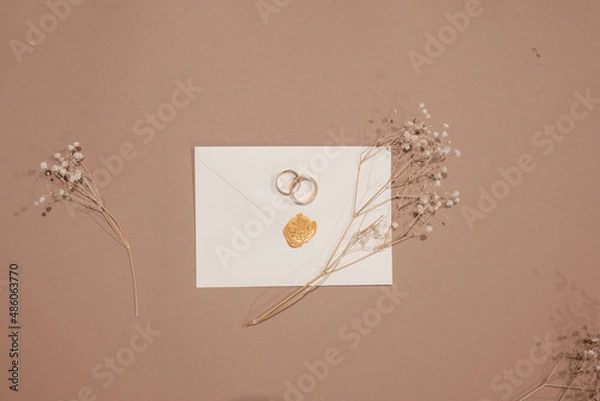 Fototapeta Wedding rings and invitations and craft envelopes on beigen background. Top view on paper cards, rings and dried flowers bouquet, copy space. Memmorable date organization concept