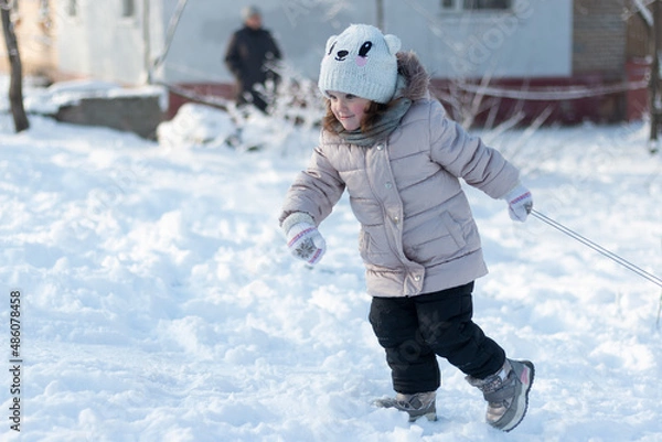 Fototapeta A diligent little child in warm clothes is struggling to pull the sled up the hill. Outdoor photo shoot on a sunny winter day