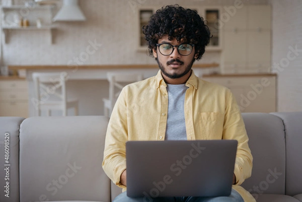 Fototapeta Pensive Indian man using laptop computer searching online looking at digital screen. Serious asian programmer working freelance project typing sitting at home. Student studying, online education