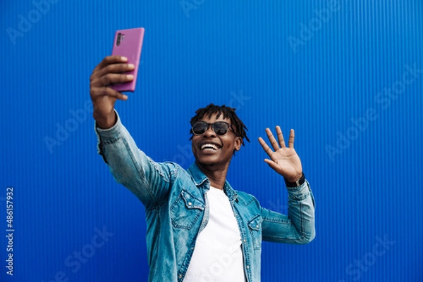 Obraz african american man in a casual shirt, on a blue background Takes a selfie on a mobile phone, making a greeting gesture