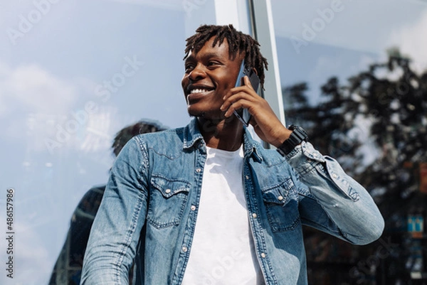 Fototapeta Image of a handsome smiling African man talking on his cell phone while walking on a city street