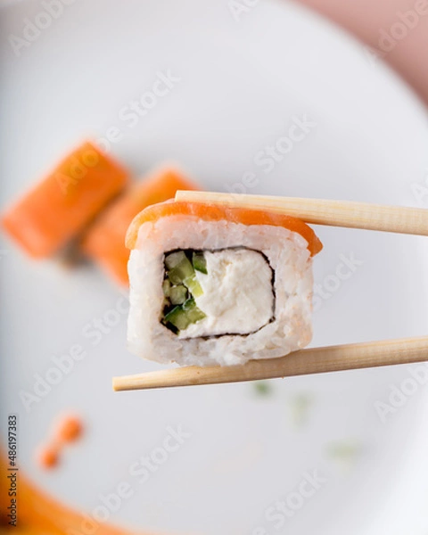 Fototapeta Salmon roll is held with chopsticks for Japanese food. In the background a plate with rolls