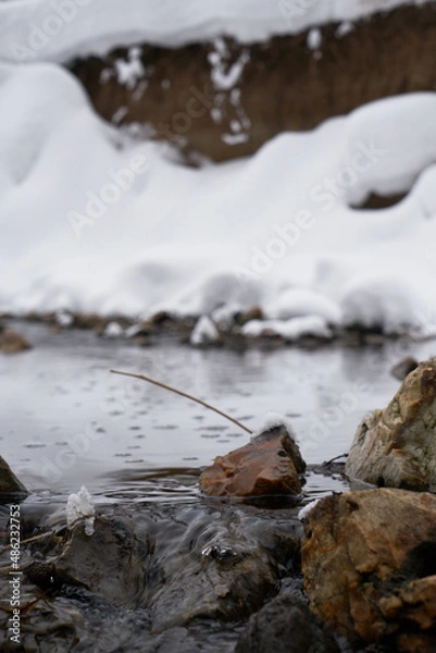 Fototapeta A small rock in the flowing river in a park. Snowy river shores. Closeup of a rock with a tiny branch sitting in the river. Open riverbed in a winter scene. in the river in a park