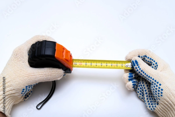 Fototapeta Hands of a worker, builder in protective gloves halding a ruler, using tape measure against white background